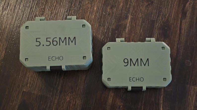 K&ouml;nnte beinhalten: Zwei olivgr&uuml;ne, rechteckige Beh&auml;lter mit Klappdeckeln. Der gr&ouml;&szlig;ere Beh&auml;lter ist mit "5.56MM" und "ECHO" beschriftet. Der kleinere Beh&auml;lter ist mit "9MM" und "ECHO" beschriftet. Beide liegen auf einer Holzoberfl&auml;che.