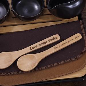 May include: Two wooden spoons with engraved messages. The first spoon reads "Love never fades". The second spoon reads "There is a reason you are Mom". The spoons are on a brown cloth with a tan cloth underneath. The spoons are on a wooden tray with black edges.