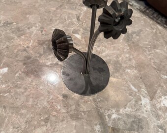 May include: A metal sculpture of three stylized flowers with gear-shaped petals. The flowers are attached to thin stems that curve upwards from a circular base. The sculpture is made of a silver-colored metal.