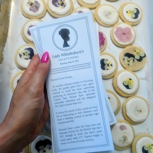 woman with pink nails holding a customizable lady whistledown pamphlet above pressed flower sugar cookies