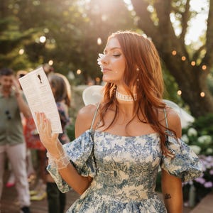 Woman dressed in Regency-inspired costume holding a Lady Whistledown Society Papers pamphlet at a garden party. Romantic outdoor setting with string lights and guests in the background, ideal for Bridgerton-themed celebrations