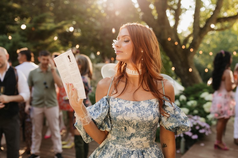 Woman dressed in Regency-inspired costume holding a Lady Whistledown Society Papers pamphlet at a garden party. Romantic outdoor setting with string lights and guests in the background, ideal for Bridgerton-themed celebrations