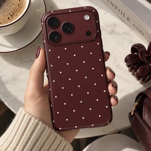Könnte beinhalten: Eine dunkelmarone Smartphone-Hülle mit einem sich wiederholenden Muster aus kleinen weißen Punkten. Die Hülle verfügt über einen erhöhten rechteckigen Kameraausschnitt mit drei kreisförmigen Linsenöffnungen und einem Blitz.