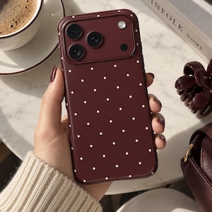 May include: A deep burgundy smartphone case featuring a repeating pattern of small white polka dots. The case has a raised rectangular camera module cutout with three circular lens openings and a circular flash opening in the upper left corner.