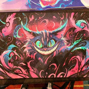 May include: A rectangular mouse mat with a colourful illustration of the Cheshire Cat from Alice in Wonderland. The design features the cat's grinning face with glowing green eyes, set against a swirling background of pink, blue, and black.