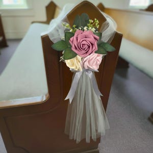 May include: A floral pew decoration featuring a cluster of three artificial roses in shades of mauve, cream, and light pink, accented with green leaves and small white filler flowers. A sheer white tulle fabric drapes downward, secured by a grey ribbon.