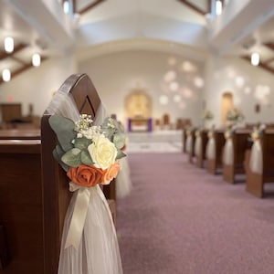 May include: A floral pew decoration featuring a cluster of artificial cream, peach, and burnt orange roses accented with green eucalyptus leaves and small white filler flowers, tied to a wooden pew with a sheer white tulle fabric and ribbon.