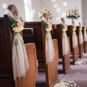 May include: A row of wooden church pews decorated with floral arrangements featuring pink and cream roses, green leaves, and white baby's breath. Each arrangement is accented with a sage green ribbon and sheer white tulle fabric draped downward.