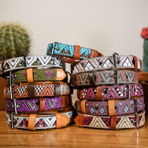 May include: A stack of colorful dog collars with geometric patterns. The collars feature various colors including turquoise, purple, orange, and white, with brown leather accents and silver buckles. Cacti in terracotta pots are in the background.