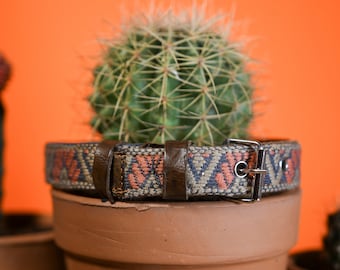 May include: A belt featuring a woven fabric strap with a repeating geometric pattern in shades of orange, blue, and tan. The belt includes a dark brown leather loop and a silver-toned metal buckle with a single prong and circular eyelets.