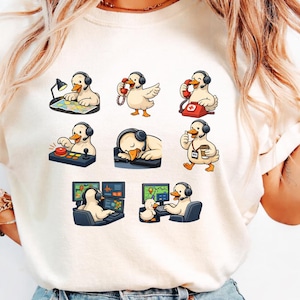 May include: Cream-colored t-shirt featuring a series of cartoon ducks wearing headsets and engaged in various activities. The ducks are depicted working at a desk, using a phone, and operating a control panel. The shirt has a crew neck.
