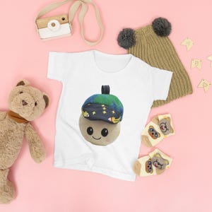 May include: A white toddler t-shirt with a playful acorn design, complete with a smiling face and a blue and green hat. The image also shows a teddy bear, a knitted hat, socks, and a toy camera, set against a pink backdrop.