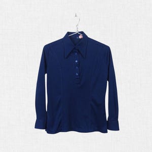 May include: A navy blue long-sleeve shirt with a pointed collar and a button-down placket. The shirt has a fitted silhouette and is hanging on a metal hanger. The fabric appears to be a smooth, knit material.