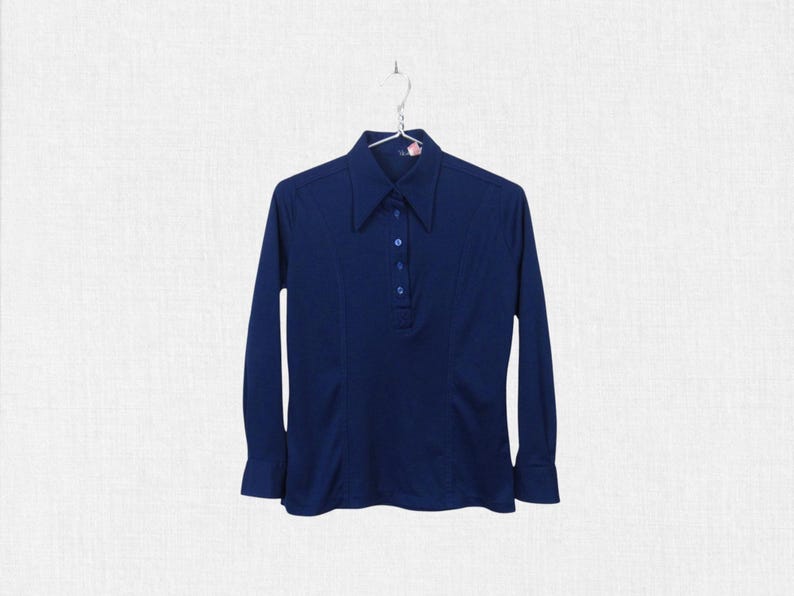 May include: A navy blue long-sleeve shirt with a pointed collar and a button-down placket. The shirt has a fitted silhouette and is hanging on a metal hanger. The fabric appears to be a smooth, knit material.