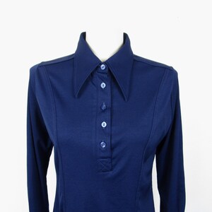 May include: A navy blue long-sleeve shirt with a large collar and button-up front. The shirt has a tailored fit and is displayed on a mannequin. The shirt is a solid color and has a classic, vintage style.
