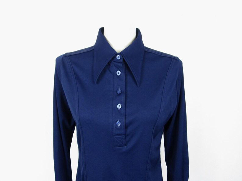 May include: A navy blue long-sleeve shirt with a large collar and button-up front. The shirt has a tailored fit and is displayed on a mannequin. The shirt is a solid color and has a classic, vintage style.