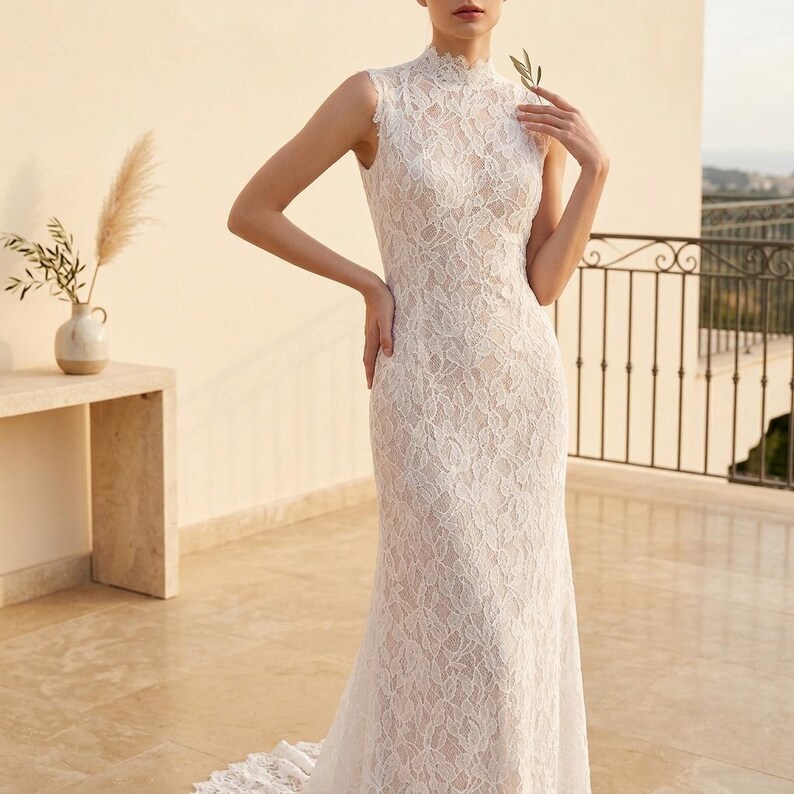May include: A sleeveless, white lace wedding dress with a high neckline and a fitted silhouette. The dress features intricate floral lace detailing and a slight train. The model is standing on a patio with a neutral background.