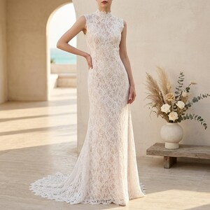 May include: An ivory lace wedding dress with a high neckline and a mermaid silhouette. The sleeveless dress features detailed floral lace and a long train. The bride is standing in a sunlit outdoor setting.