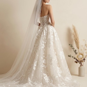 May include: A strapless ivory wedding gown with floral lace appliques and a long, flowing skirt. The back features a corset-style closure. A sheer veil cascades down the back. The dress is displayed on a person.