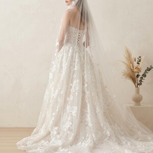 May include: A strapless ivory wedding dress with floral lace detailing and a long train. The back features a corset design. A sheer veil cascades down the back, adding to the elegant bridal look.