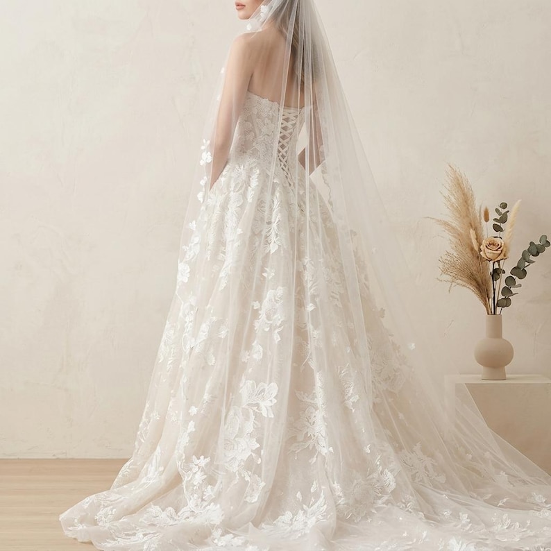May include: A strapless ivory wedding dress with floral lace detailing and a long train. The back features a corset design. A sheer veil cascades down the back, adding to the elegant bridal look.