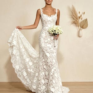 Puede incluir: Vestido de novia de encaje blanco con corpiño ajustado y falda fluida. El vestido presenta un estampado floral y un dobladillo festoneado. La novia sostiene un ramo de flores blancas.