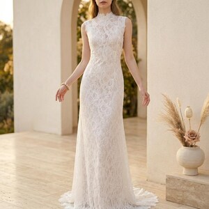 May include: A sleeveless, floor-length white lace wedding dress with a high neckline. The dress features intricate floral lace detailing and a fitted silhouette that flares out at the bottom. The bride is wearing a bracelet.