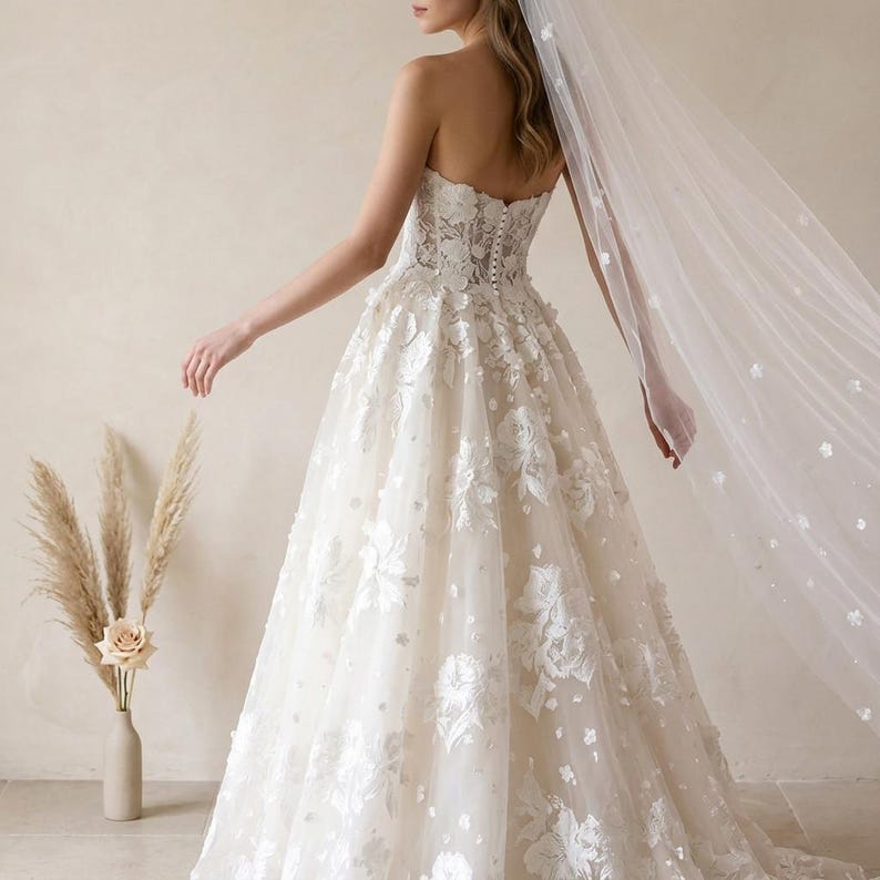 May include: A strapless ivory wedding gown with floral appliqu&eacute;s and a flowing skirt. The bodice features intricate lace detailing, and a sheer veil with delicate floral accents cascades down. A bouquet of dried flowers is in a vase.
