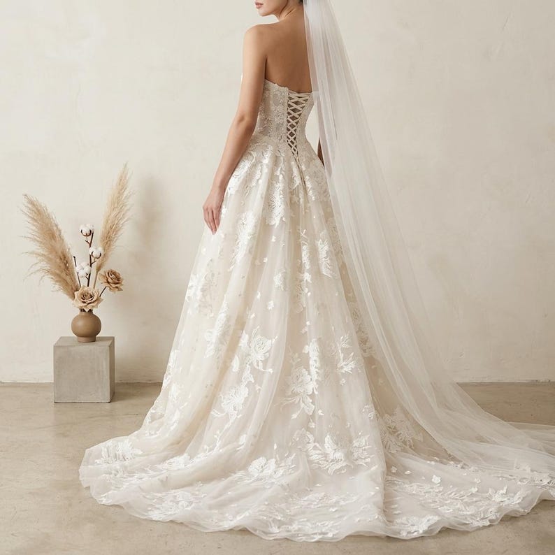 May include: A strapless ivory wedding gown with intricate floral lace detailing. The dress features a corset back and a flowing skirt that extends into a long train. A sheer veil cascades down the back, adding to the elegant bridal look.