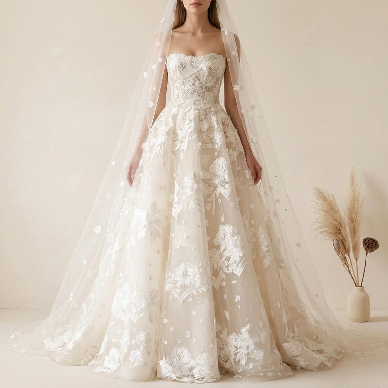 May include: A strapless ivory wedding gown with floral embroidery and a flowing skirt. The dress is paired with a sheer veil adorned with small, scattered embellishments. The gown's bodice features intricate detailing.