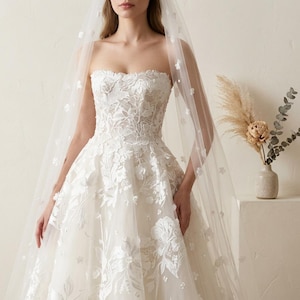 May include: A strapless white wedding dress with floral lace detailing. The dress features a full skirt and is paired with a sheer veil adorned with small, delicate flowers. The veil cascades down the back, adding a touch of elegance.