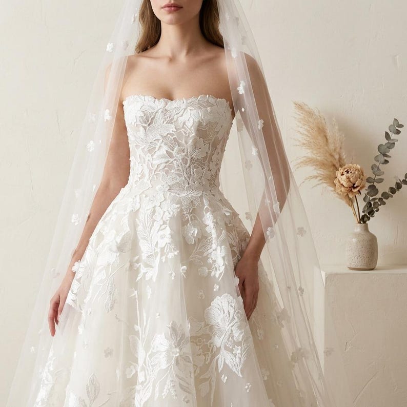 May include: A strapless white wedding dress with floral lace detailing. The dress features a full skirt and is paired with a sheer veil adorned with small, delicate flowers. The veil cascades down the back, adding a touch of elegance.
