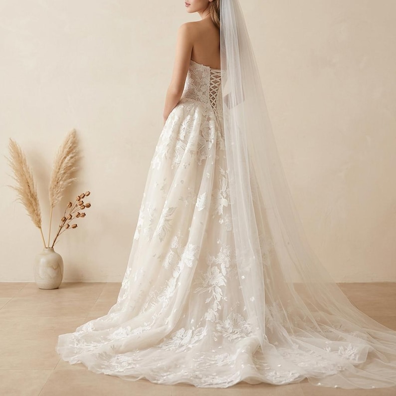 May include: A strapless ivory wedding dress with floral lace detailing and a flowing skirt. The back features a corset design. A sheer veil cascades down the back, adding to the elegant bridal look.