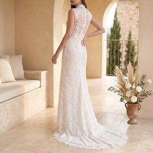 May include: A white lace wedding dress with a high neckline and a long train. The sleeveless dress is fitted through the bodice and hips, then flares out at the hem. The dress is made of delicate lace and has an elegant design. The dress is a classic bridal gown.