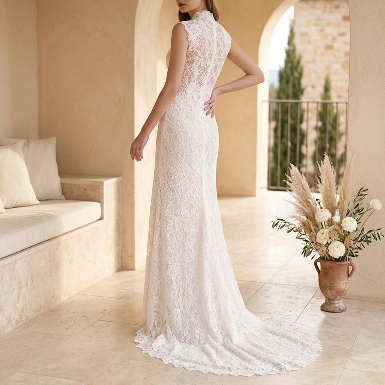 May include: A white lace wedding dress with a high neckline and a long train. The sleeveless dress is fitted through the bodice and hips, then flares out at the hem. The dress is made of delicate lace and has an elegant design. The dress is a classic bridal gown.