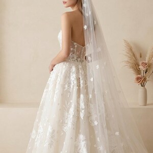 May include: A strapless ivory wedding dress with floral embroidery and a matching veil. The veil is adorned with delicate flower appliqu&eacute;s. The dress features a fitted bodice and a full skirt, creating an elegant silhouette.