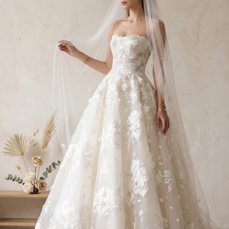 May include: A strapless ivory wedding gown with floral lace appliques and a full skirt. The dress is paired with a sheer veil adorned with small embellishments. Gold bracelets are visible on the model's wrists.