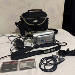 May include: A silver Samsung camcorder with a black strap and a black carrying case. The strap has the word "SAMSUNG" printed on it. Also included are two miniDV tapes, a power adapter, and cables.