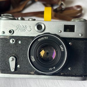 May include: A vintage silver and black FED 3 camera with a black lens. The camera has the text "FED 3" in Cyrillic script. The lens has the numbers "8658751" and "2.8/52" printed on it. A yellow strap is attached.