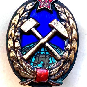 May include: A gold-colored metal badge with a red star and hammer and sickle emblem at the top. The badge features two crossed hammers over a globe, surrounded by a wreath of leaves. The globe is blue and green, and the background is blue.