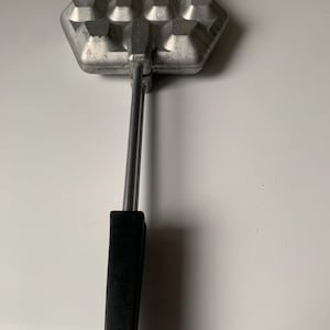 May include: A silver waffle iron with a hexagonal shape and a black handle. The iron has a waffle pattern with 12 squares.