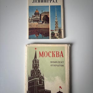 Op de afbeelding: Twee vintage reisgidsen. Het bovenste boek heeft de titel "LENINGRAD" met afbeeldingen van gebouwen en een blauw-witte omslag. Het onderste boek heeft de titel "MOSKVA" met een rode ster en een illustratie van een klokkentoren op een groene omslag.