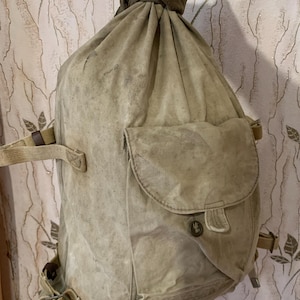 May include: A large, tan canvas bag with a drawstring closure and a large flap pocket on the front. The bag has a worn, vintage look and is made of heavy-duty canvas.