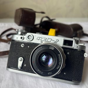 Puede incluir: Una cámara FED-2 vintage en blanco y negro con una funda de cuero. La cámara tiene una lente con el texto "ИНДУСТАР-26 м" y "F=5CM" impreso. La cámara está sobre una superficie blanca.
