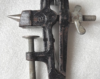 May include: A small, vintage black metal vise with a silver-colored screw and handle. The vise has a pointed anvil on one side and a screw mechanism for clamping objects. The vise is likely used for holding small items during crafting or repair.