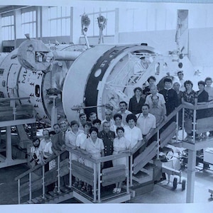 以下が含まれることがあります： 大きな円筒形の機械の前に立っている人々の白黒写真。機械は金属製のリングがいくつかあり、工場や産業施設の一部であるように見える。