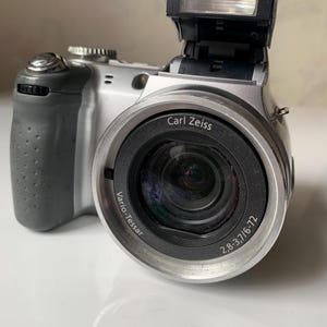 Puede incluir: Una cámara digital plateada con una lente Carl Zeiss Vario-Tessar. La lente tiene un rango de zoom de 2.8-3.7/6.7-72. La cámara tiene un flash incorporado.