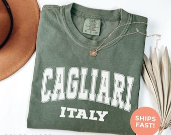 May include: Moss green t-shirt with the word "CAGLIARI" in white block letters above "ITALY". A gold necklace rests on the shirt. The shirt has a Comfort Colors tag. A brown hat and dried leaves are in the background.