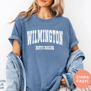 May include: Blue jean colored t-shirt with the word "WILMINGTON" in white block letters above "NORTH CAROLINA". The shirt has rolled-up sleeves and is worn with a denim jacket. A round orange sticker says "SHIPS FAST!"