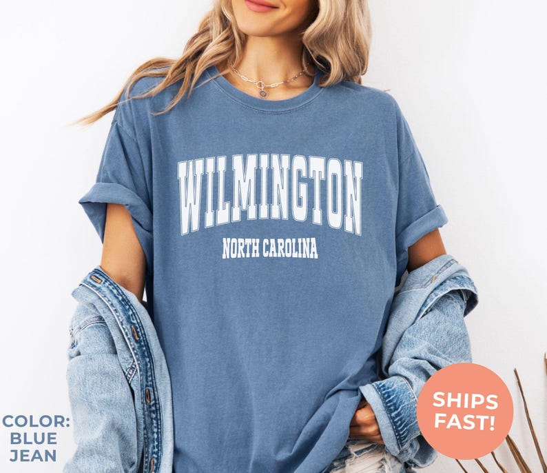 May include: Blue jean colored t-shirt with the word "WILMINGTON" in white block letters above "NORTH CAROLINA". The shirt has rolled-up sleeves and is worn with a denim jacket. A round orange sticker says "SHIPS FAST!"